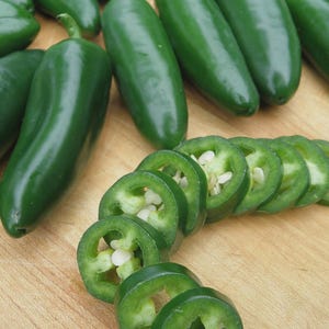 May include: Fresh, vibrant green jalapeño peppers, some whole and others sliced, revealing white seeds. The peppers are arranged on a light brown wooden surface, showcasing their smooth texture and bright color.