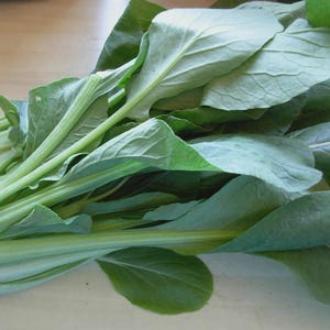May include: A close-up of fresh green leafy vegetables, likely bok choy or a similar Asian green. The leaves are a vibrant green with visible veins, and the stems are a lighter green. The vegetables are arranged on a light-colored surface.