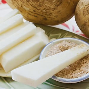 May include: A close-up of jicama, a root vegetable, cut into sticks and served with a small bowl of seasoning. Two whole jicama bulbs are in the background. The jicama sticks are white and the seasoning is brown.