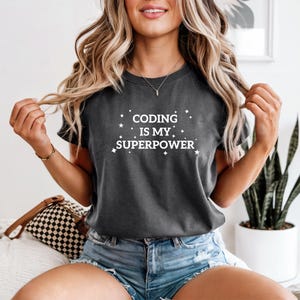 May include: Dark grey t-shirt with the white text "CODING IS MY SUPERPOWER". The text is surrounded by small star and diamond shapes. The shirt is a crew neck and has short sleeves.