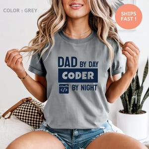 Op de afbeelding: Een grijs t-shirt met de tekst "DAD by day CODER by night" in marineblauw. Het shirt heeft een ronde hals en korte mouwen. De tekst bevat een computercode-pictogram.