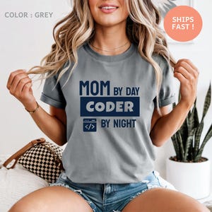 May include: Grey t-shirt with the text "MOM by DAY CODER by NIGHT" in navy blue. The shirt features a computer code icon. The shirt is a casual wear item.