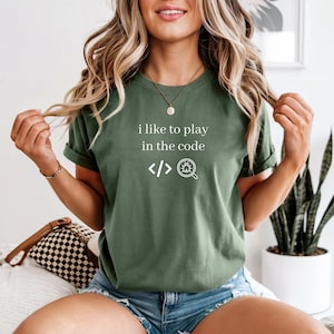 May include: Olive green t-shirt with the text "i like to play in the code" and code symbols. The shirt has short sleeves and a relaxed fit. The design is in white. The shirt is made of a soft material.