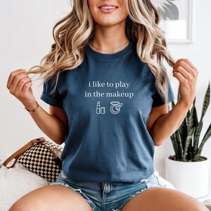 Op de afbeelding: Marineblauw T-shirt met de witte tekst "i like to play in the makeup". Daaronder twee witte make-up iconen: een lippenstift en een make-up compact. Het shirt is gemaakt van een zacht materiaal.