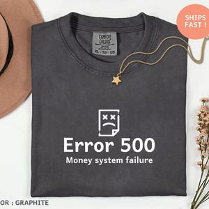 Op de afbeelding: Grafietkleurig T-shirt met de witte tekst "Error 500 Money system failure". Het shirt heeft een ronde hals en korte mouwen. Een gouden ketting met een sterhanger ligt op het shirt.