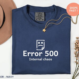 May include: Navy blue t-shirt with the text "Error 500 Internal chaos" in white. The shirt features a graphic of a sad document icon. A gold star pendant necklace rests on the shirt. The shirt is labeled "Comfort Colors" and is size XL-TO-EG.