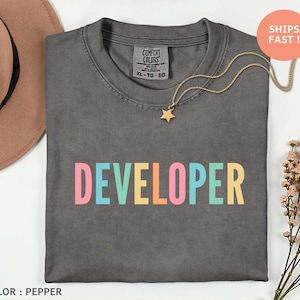 May include: A dark gray t-shirt with the word "DEVELOPER" printed in colorful letters. The shirt has a crew neck and short sleeves. A gold necklace with a star pendant is draped across the shirt.
