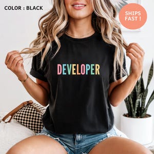 A woman with long, wavy blonde hair wearing a black t-shirt with the word "Developer" printed on it. She is posing with her hands behind her back, and there is a potted plant visible in the background.
