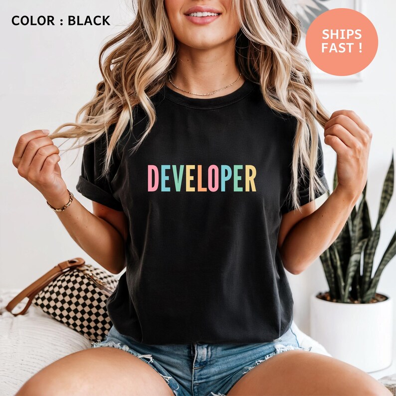A woman with long, wavy blonde hair wearing a black t-shirt with the word "Developer" printed on it. She is posing with her hands behind her back, and there is a potted plant visible in the background.