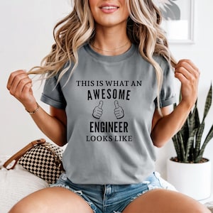 Könnte beinhalten: Graues kurzärmeliges T-Shirt mit dem schwarzen Text "THIS IS WHAT AN AWESOME ENGINEER LOOKS LIKE". Das Design enthält zwei Daumen-hoch-Symbole. Das Shirt ist aus weichem Material.