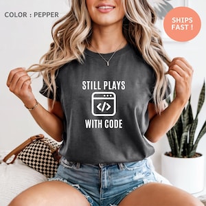 May include: Dark grey t-shirt with the text "STILL PLAYS WITH CODE" and a graphic of a computer screen with code symbols. The shirt is a pepper colour.