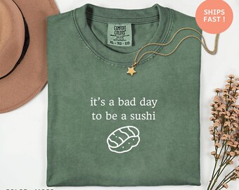 May include: A moss green t-shirt featuring the phrase "it's a bad day to be a sushi" and a sushi illustration. The shirt has a crew neck and short sleeves. A gold necklace with a star pendant is on the shirt.