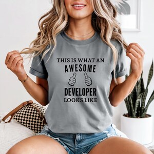 Puede incluir: Una camiseta gris con el texto "THIS IS WHAT AN AWESOME DEVELOPER LOOKS LIKE" en negro. El diseño incluye dos símbolos de pulgares arriba. La camiseta es de cuello redondo y manga corta.