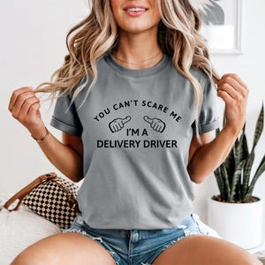 May include: A gray t-shirt with the text "YOU CAN'T SCARE ME I'M A DELIVERY DRIVER" in black. The shirt has short sleeves and a crew neck. The person is wearing blue denim shorts.