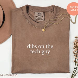 May include: Espresso-colored t-shirt with the text "dibs on the tech guy." The shirt has a crew neck and a Comfort Colors tag. A gold necklace with a star pendant is draped across the shirt. The image also includes a hat and flowers.