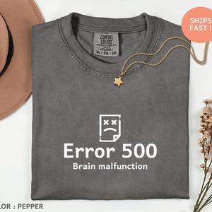 May include: Pepper-colored t-shirt with the text "Error 500 Brain malfunction" and a graphic of a sad face on a document. The shirt has a crew neck and a Comfort Colors tag. A gold necklace with a star pendant is on the shirt.