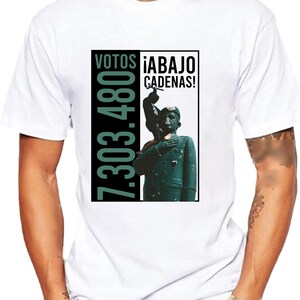 May include: White t-shirt with a black and green graphic print. The graphic features a person holding a hammer and the text "VOTOS 7.303.480 &iexcl;ABAJO CADENAS!"