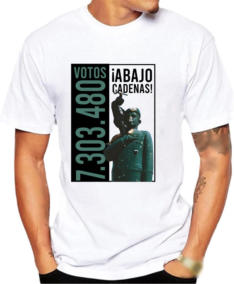 May include: White t-shirt with a black and green graphic print. The graphic features a person holding a hammer and the text "VOTOS 7.303.480 &iexcl;ABAJO CADENAS!"