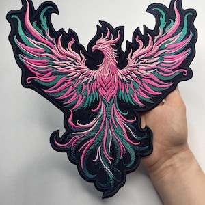May include: Embroidered patch featuring a phoenix design with outstretched wings. The patch showcases a color palette of pink, teal, and black. The phoenix is intricately detailed, with a black border. This patch is suitable for jackets, bags, or other fabric items.