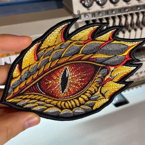 May include: An embroidered patch featuring a detailed dragon eye with a vertical black pupil, surrounded by a vibrant red iris and textured scales in shades of metallic gold, yellow, and grey, all outlined with a thick black border.