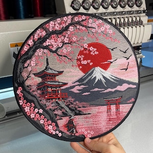 May include: A circular embroidered patch featuring a landscape with a red sun, a snow-capped mountain, a pagoda, a torii gate in water, and a cherry blossom tree with pink flowers. The design includes a small figure in the foreground.