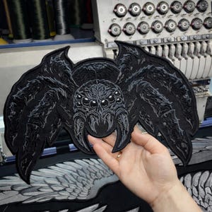 May include: A large, black embroidered spider patch with detailed wings and multiple eyes. The patch is held up in front of an embroidery machine, showcasing the intricate stitching and design. The spider's body and legs are textured.
