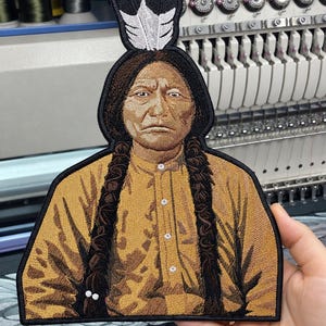 May include: An embroidered patch featuring a portrait of Sitting Bull with long dark braided hair and a feather headdress. The figure wears a tan button-down shirt. The patch has a black border and is displayed against an embroidery machine.