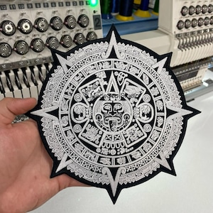Peut inclure: Un écusson brodé noir et blanc représentant un calendrier aztèque détaillé. L'écusson a une forme d'étoile avec des bords pointus. Le texte "Made Soul Studio" est visible sur l'écusson. L'écusson est tenu devant une machine à broder.