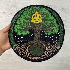 Op de afbeelding: Ronde zwarte patch met een geborduurd Tree of Life-ontwerp. De boom heeft groene bladeren, een bruine stam en ingewikkelde wortels. Een gouden Keltische knoop staat bovenaan, met een groene rand en sterren.