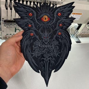 Peut inclure: Un grand écusson brodé noir avec un motif gothique détaillé. L'écusson représente une figure démoniaque avec de multiples yeux rouges, des ailes et une épée. L'œil central est orange. L'écusson est tenu dans une main.