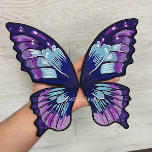 May include: Embroidered butterfly wings in shades of purple, blue, and white. The wings feature intricate stitching and a detailed design. The wings are a craft supply, perfect for adding a whimsical touch to clothing or accessories.