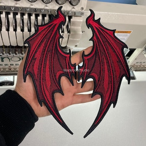May include: Pair of embroidered red and black devil wings. The wings are detailed with a textured pattern and a black outline. The wings are held in a person's hand, with the embroidery machine in the background.
