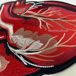 May include: Embroidered patch depicting a detailed anatomical heart. The heart is predominantly red, with varying shades, and features white, brown, and black embroidery. The text "MadeSoulStudio" is visible.