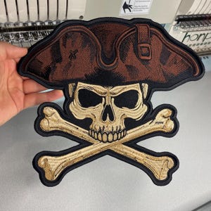 May include: A large embroidered patch featuring a brown pirate hat worn over a beige human skull with black eye sockets and nose cavity, positioned above two crossed beige bones, all outlined with a thick black border on a black fabric base.