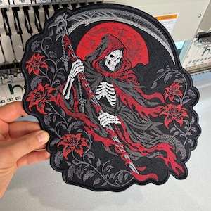 May include: A large circular embroidered patch featuring a skeleton figure in a black and red hooded cloak holding a scythe. The design includes a large red moon in the background and red lily flowers with grey vines surrounding the central figure.