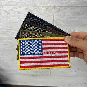 Könnte beinhalten: Gestickte Aufnäher mit der amerikanischen Flagge in verschiedenen Farbschemata. Der Hauptaufnäher zeigt die Flagge mit roten und weißen Streifen, einem blauen Sternenfeld und einem gelben Rand. Weitere Aufnäher zeigen Tarn- und monochrome Designs.