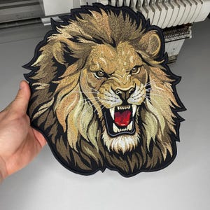 May include: Embroidered lion head patch with a fierce expression. The patch features detailed stitching in shades of brown, tan, and gold, with a black border. The lion's mouth is open, revealing red teeth and a red tongue. This patch is suitable for jackets, bags, or other apparel.