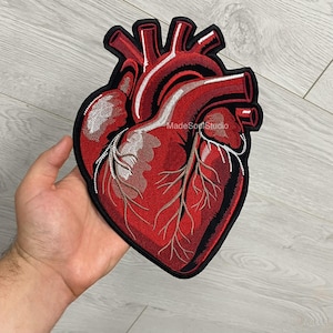 May include: Embroidered patch in the shape of a human heart. The patch features a detailed design with red, black, and silver thread. The text "MadeSonStudio" is visible on the patch. The patch is held in a hand.