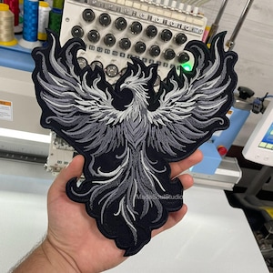 Może przedstawiać: Haftowana naszywka z motywem feniksa w odcieniach szarości i bieli na czarnym tle. Naszywka ma skomplikowane detale, ze skrzydłami i piórami ogona feniksa rozłożonymi szeroko. Naszywka jest trzymana w dłoni osoby.