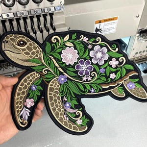 May include: An embroidered patch shaped like a sea turtle with a black border. The turtle head and flippers feature tan and gold scale patterns. The shell is decorated with intricate green leaves and various purple and pink floral embroidery.