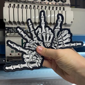 May include: Three embroidered patches featuring white skeleton hands on a black background. The patches display different hand gestures, including a middle finger, a peace sign, and a shaka sign, all rendered in detailed white thread.