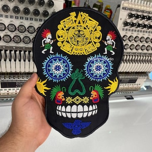 May include: Black skull-shaped embroidered patch with colourful Aztec-inspired designs. Features a central sun design, blue starburst eyes, and a green heart-shaped nose. Includes the text "MadeSoulStudio".