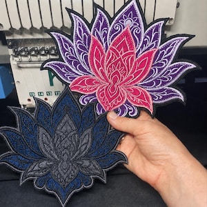 May include: Two embroidered lotus flower patches. The top patch features vibrant pink and purple thread with intricate white filigree detailing. The bottom patch displays a dark blue and charcoal grey colour scheme with similar ornate stitching.