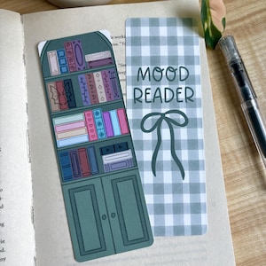 May include: Two rectangular bookmarks. One bookmark features a dark green bookshelf illustration filled with colorful books, including some with heart and wing motifs. The second bookmark has a sage green gingham pattern with the text MOOD READER.