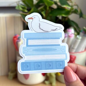 May include: A die-cut sticker featuring a white seagull with a closed eye resting on a stack of four light blue books. The bottom book displays a repeating pattern of four square icons, each containing a small seashell illustration.