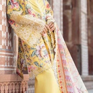 Op de afbeelding: Een gele outfit met bloemenprint, bestaande uit een tuniek met lange mouwen en een bijpassende broek. De outfit heeft gedetailleerde kant- en bloempatronen, met een doorschijnende, bedrukte sjaal. Het kleurenpalet omvat geel, roze en wit.