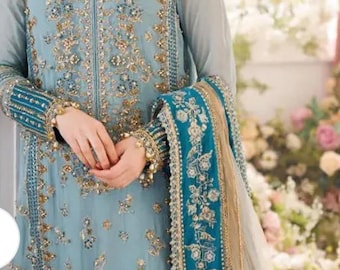 Puede incluir: Vestido bordado azul claro con detalles dorados y turquesas. El vestido presenta intrincados diseños florales y adornos. Las mangas y los bordes están adornados con tela turquesa. Un collar y una pulsera dorados completan el look.