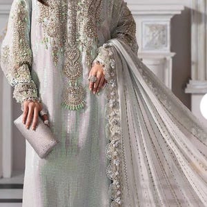 Może przedstawiać: Jasna, zdobiona suknia wieczorowa z misternymi zdobieniami z koralików i cekinów. Suknia ma długie rękawy, wysoki dekolt i pasujący szal. W dłoni trzymana jest srebrna kopertówka. Odpowiednia na specjalne okazje.