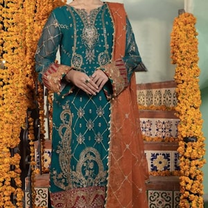 May include: Teal embroidered formal dress with gold accents and a rust-colored dupatta. The dress features intricate detailing and is worn with matching teal pants. The background includes marigold garlands and decorative tile.