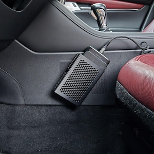 May include: A black rectangular electronic device with a honeycomb-patterned front grille, mounted to the side of a car center console. The device is connected by a black coiled cable and features a textured top surface with a small logo.